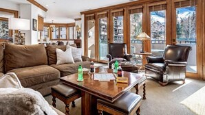 A spacious living room with a brown leather sofa, a wooden coffee table, and a view of a mountain landscape through large windows. - Ski in, Ski out 3 Bedroom Condo at the Beaver Creek Lodge (Avon)