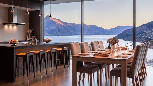 A modern dining area with a large table and chairs, a bar with stools, and a view of mountains and a lake. - The Belfast (Queenstown)