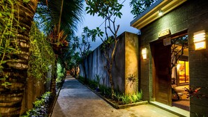 A narrow pathway flanked by lush greenery and a building with a modern facade and illuminated signage.