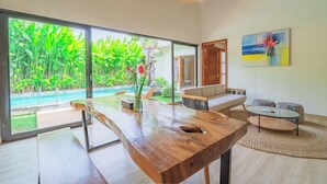 A modern living room with a wooden dining table, a sofa, and a coffee table. There is a large window with a view of a swimming pool and lush greenery.