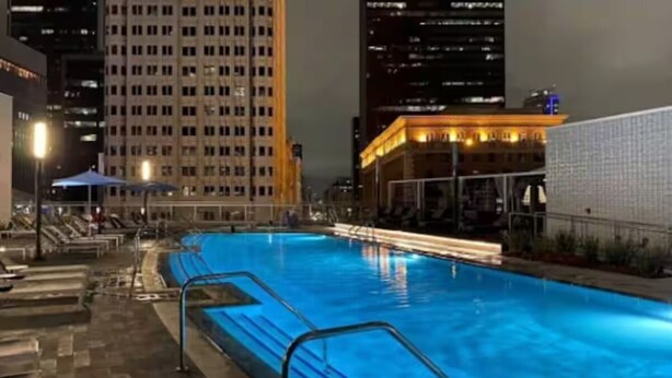 A swimming pool with a tiled deck and lounge chairs, surrounded by high-rise buildings at night.