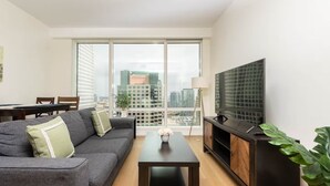 A modern living room with a grey sofa, a coffee table, a TV on a wooden cabinet, and a city view through large windows.