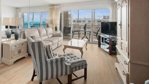 Un salon spacieux avec un grand canapé, un fauteuil à rayures, une table basse en verre et un téléviseur. La pièce est dotée de grandes fenêtres qui offrent une vue sur la ville.