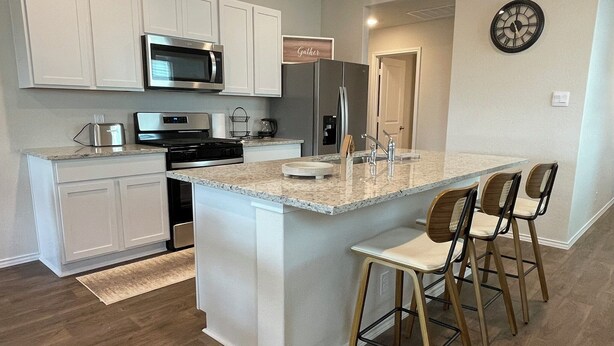 Una cocina moderna con armarios blancos, una isla central con taburetes altos, electrodomésticos de acero inoxidable y un reloj en la pared.