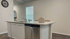 Una cocina con muebles blancos, lavavajillas, encimera de granito y un reloj en la pared.