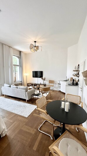 A modern living room with a sofa, chairs, a dining table, and a TV. - Orangerie · Frauenapartment mit Gartenbalkon (Kassel)