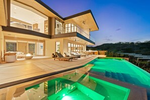 A modern two-story house with a large glass-enclosed balcony, a swimming pool, and a well-lit interior. - Villa Zaya - Designer Ocean-View Villa in Reserva Conchal (Santa Cruz)