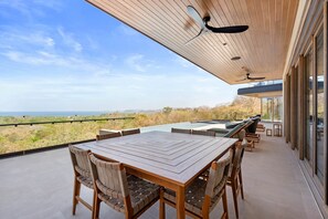 A spacious outdoor patio with a wooden table and chairs, a ceiling fan, and a view of the landscape.