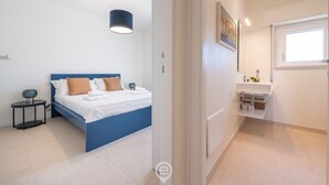 Et moderne soveværelse med en blå seng, hvide vægge, et lille natbord og en hængende lampe.