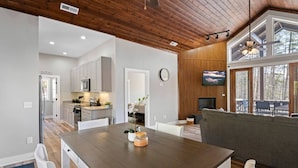A modern kitchen with white cabinets, a dining table with chairs, and a living area with a gray sofa and a TV.