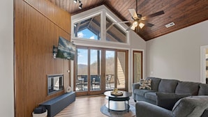 A spacious living room with a high ceiling, wooden paneling, a fireplace, a television, and a large sliding glass door leading to an outdoor patio.