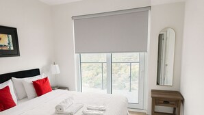 A neatly arranged bedroom with a large bed, a bedside table, a nightstand, a window with a view, and a mirror.