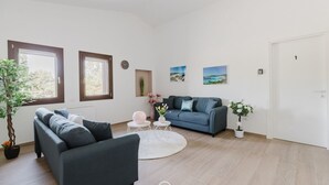 A modern living room with a grey sofa, a blue sofa, and a round coffee table. There are two windows with brown frames, a white door, and a clock on the wall. - House Adele - Dreaming Vacation (Villa San Pietro)