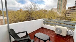 Un balcon abrité avec deux chaises noires et une petite table, un climatiseur blanc et une vue sur des immeubles et des arbres.