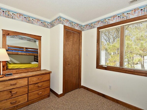 A bedroom with a wooden dresser, a bed with a headboard mirror, a window with blinds, and a door.