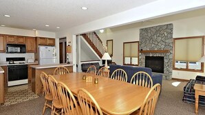 Una cocina amplia con armarios de madera, un comedor con una mesa grande y sillas de madera, una escalera que sube a un altillo, y una chimenea de piedra.