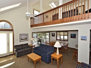 A spacious living area with a high ceiling, a wooden balcony, a dining area with chairs and a table, and a sofa.