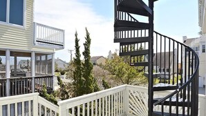 Una escalera de caracol con barandilla blanca, un espacio exterior cubierto con terraza de madera y un edificio residencial al fondo.