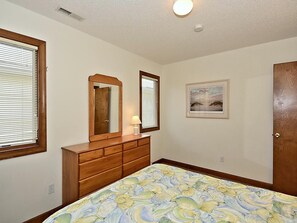 A bedroom with a wooden dresser, a bed with a patterned bedspread, a wall-mounted mirror, a framed picture, and a door.