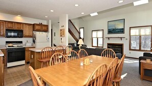 A spacious living area with a wooden dining table, chairs, a kitchen with wooden cabinets, a staircase, and a large window with a view.