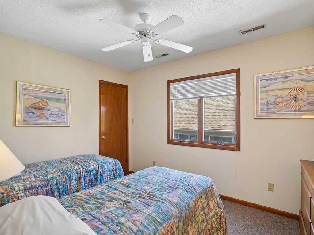 A bedroom with two beds, a ceiling fan, a window with blinds, and artwork on the walls.