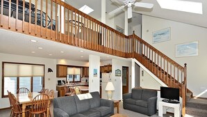 A spacious living area with a dining table, two sofas, a coffee table, and a TV stand. A staircase with a wooden railing leads to an upper level.