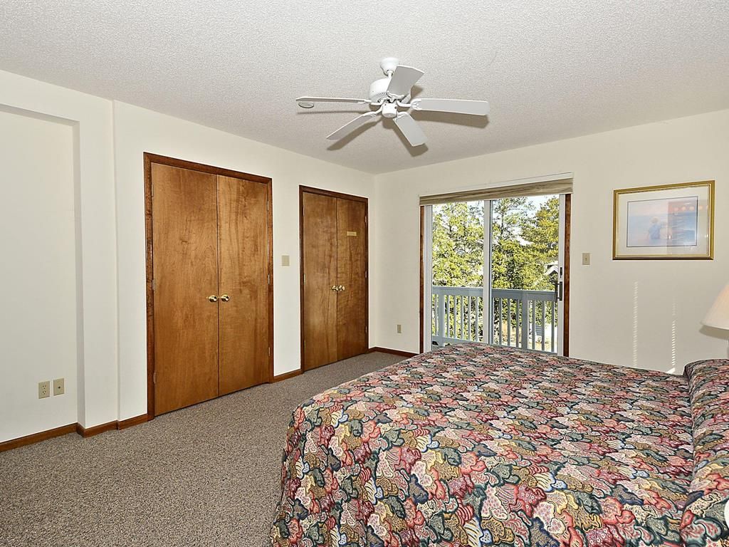 A bedroom with a bed, two wooden doors, a ceiling fan, a window with a view, and a framed picture on the wall.