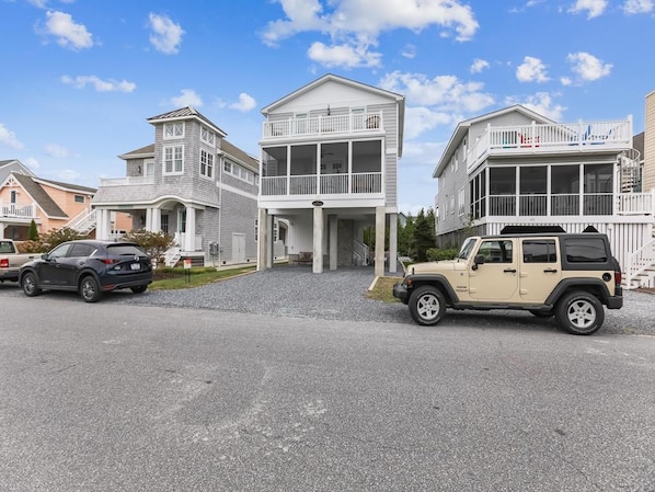 A residential area with a two-story house featuring a balcony, a driveway with two cars, and a clear sky. - Oceanview Beach House w/ Outdoor Lounges (Bethany Beach)