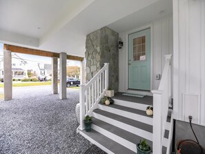 A covered porch with a stone wall, a teal door, and a staircase with potted plants. - Oceanview Beach House w/ Outdoor Lounges (Bethany Beach)