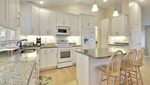 A modern kitchen with white cabinets, a granite countertop, a microwave, an oven, and a refrigerator. There are three wooden bar stools at the island. - 6-Bedroom Beach House w/ Ocean Views (South Bethany Beach)