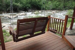 Eine hölzerne Schaukelbank auf einer Holzterrasse mit Blick auf einen Fluss, Felsen und Grünflächen.