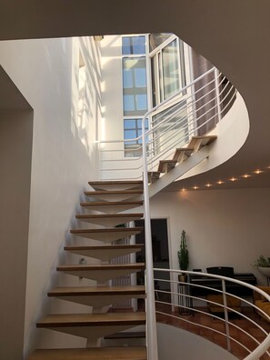 Una escalera en espiral con barandilla blanca y escalones de madera, que lleva a un balcón con vistas al cielo y edificios.