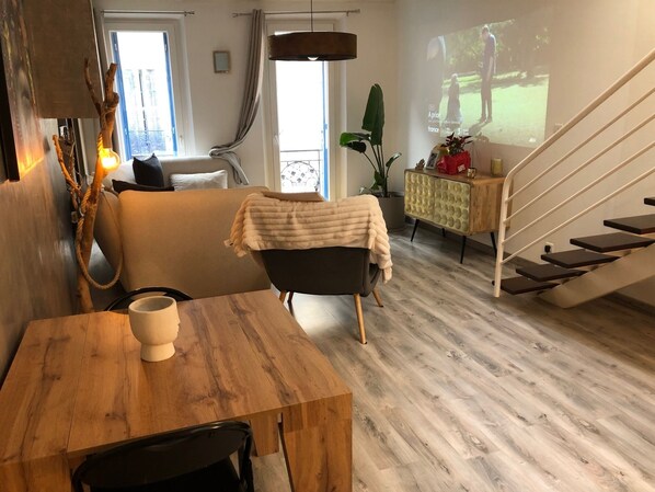 Una sala de estar moderna con una mesa de comedor de madera, un sofá, un sillón negro, una escalera y un ventanal con vistas a un parque.