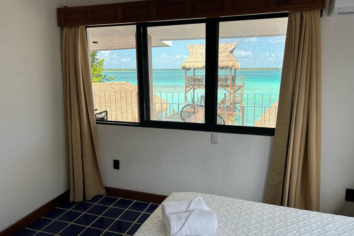 Une chambre d’hôtel avec un lit, une fenêtre avec des rideaux et une vue sur une plage bordée de parasols en chaume.