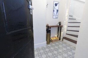 A hallway with patterned flooring, a staircase, a bench, a trash bin, and a wall-mounted artwork. - Lovely 2-Bed Town Centre Duplex Apartment (Gloucestershire)