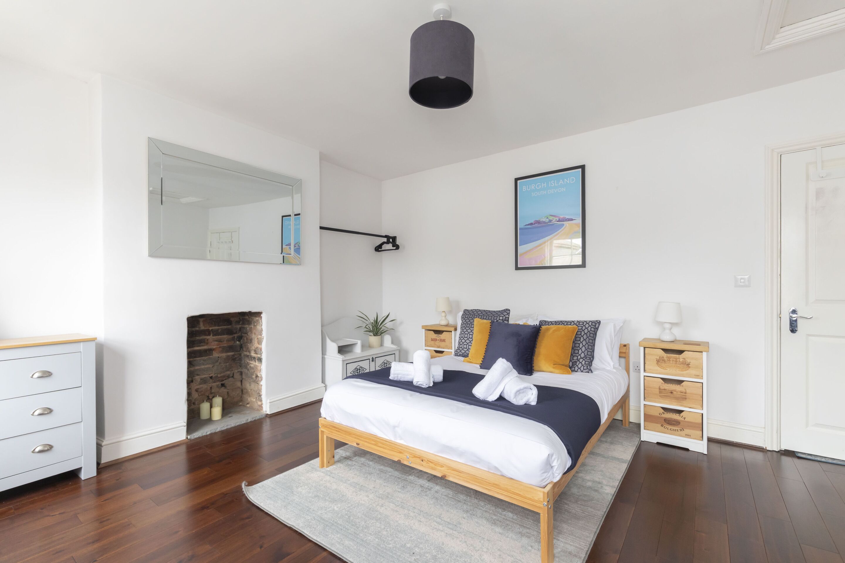 A modern bedroom with a wooden bed, a white dresser, a mirror, a fireplace, and a framed picture on the wall.