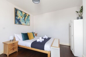 En pænt redt seng med hvide sengetøj, et mørkeblåt tæppe og en træsenggavl. Et lille natbord i træ har en lampe og en skuffe. Et stort abstrakt maleri hænger på væggen over sengen.