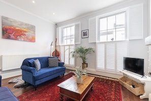 A living room with a blue sofa, a wooden coffee table, a red patterned rug, a potted plant, a guitar, and a television.