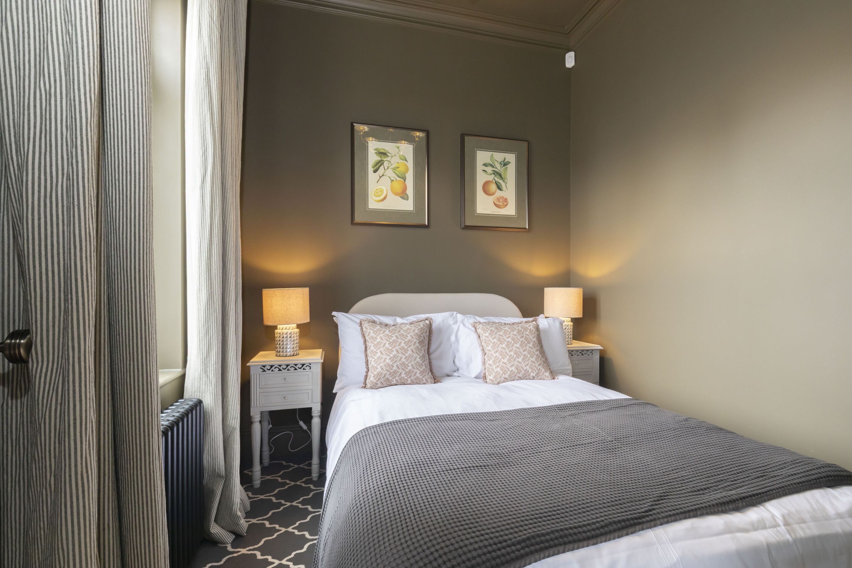 A hotel room with a bed, two bedside lamps, a radiator, a window with curtains, and two framed botanical prints on the wall.
