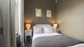 A hotel room with a bed, two bedside lamps, a radiator, a window with curtains, and two framed botanical prints on the wall.