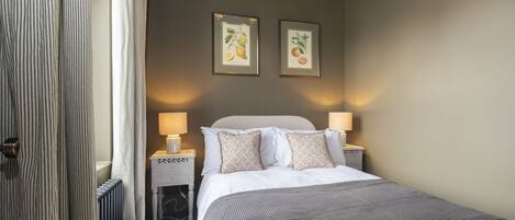A hotel room with a bed, two bedside lamps, a radiator, a window with curtains, and two framed botanical prints on the wall.