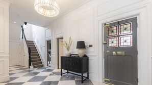 Un couloir doté d’un sol à carreaux, d’un escalier, d’une console noire surmontée d’un vase et d’une porte ornée de panneaux de vitrail.