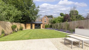 A backyard with a lawn, a wooden fence, a house in the background, and a patio area with a table and chairs. - Luxury 6 Bedroom Period Home with Sauna and Gym (Gloucestershire)