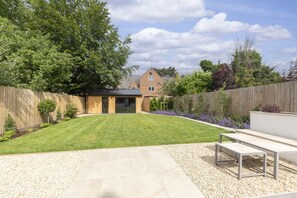 A backyard with a lawn, a wooden fence, a house in the background, and a patio area with a table and chairs. - Luxury 6 Bedroom Period Home with Sauna and Gym (Gloucestershire)