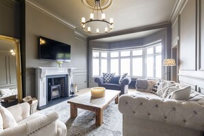 A living room with a fireplace, a television mounted on the wall, and a large window providing natural light. - Luxury 6 Bedroom Period Home with Sauna and Gym (Gloucestershire)