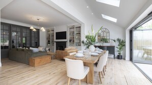 Un vaste espace de vie comprenant une table à manger en bois, des chaises blanches et un canapé. On y trouve également une cheminée, des étagères ornées d’objets décoratifs et une grande fenêtre qui laisse entrer la lumière naturelle.