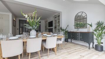 A dining area with a wooden table set for four, surrounded by chairs. There is a large vase with flowers on the table, and a dark-colored cabinet with drawers to the right.