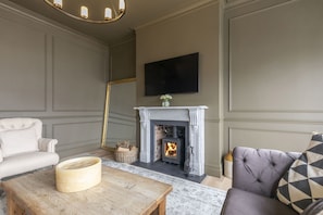 A living room with a fireplace, a sofa, a coffee table, and a television mounted on the wall. - Luxury 6 Bedroom Period Home with Sauna and Gym (Gloucestershire)