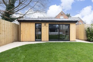 A wooden structure with a glass door, likely a shed or storage unit, in a backyard with a lawn. - Luxury 6 Bedroom Period Home with Sauna and Gym (Gloucestershire)