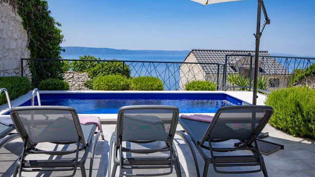 Ein Poolbereich mit zwei Liegestühlen und einem großen Sonnenschirm, mit Blick auf das klare blaue Meer und sanfte Hügel in der Ferne.